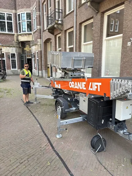 Aanhangerlift van Verhuislifthuren.nl opgesteld op straat bij bakstenen pand, verhuizer erbij. Familiebedrijf dat eerlijk zaken doet tegen een vaste prijs.