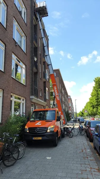 Oranje Verhuislifthuren.nl-vrachtwagen met ladder omhoog langs bakstenen flat met balkons. Onze ladderlift hijst zware kasten zo de hoogte in.