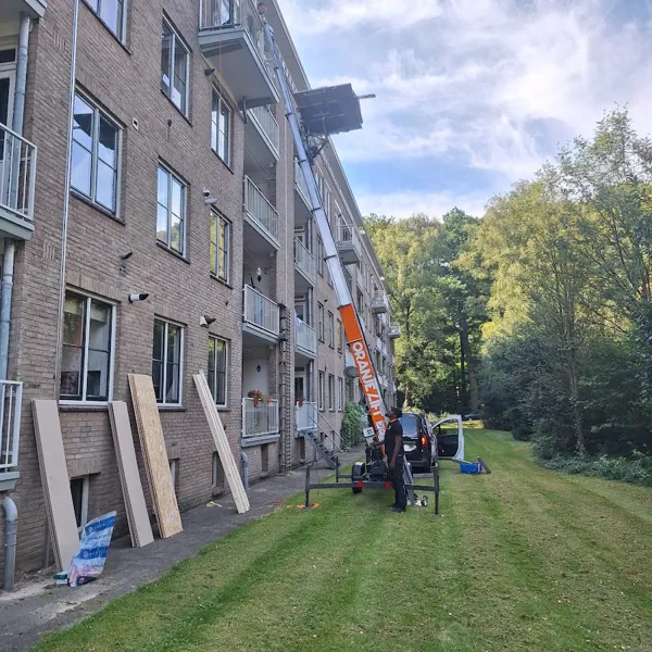 Op het gras naast een bakstenen flat, aanhangerlift met ladder tot bovenste balkon. Bij Verhuislifthuren.nl huur je een verhuislift altijd met operator.