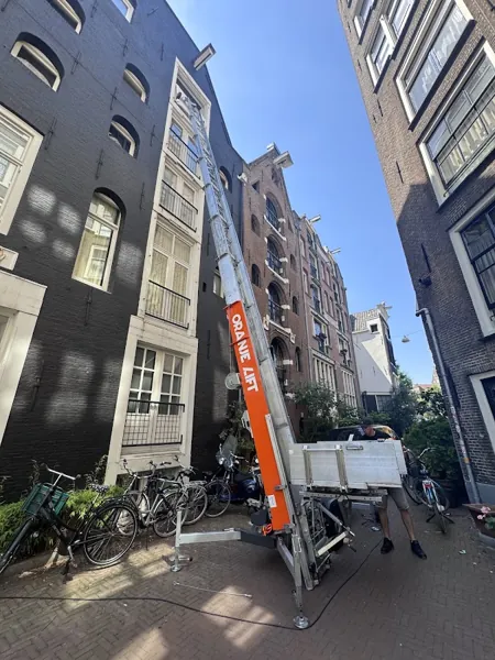 Smalle straat, aanhangerlift met ladder naar bovenste raam van zwart geverfd pand. Vandaag gebeld, morgen staat Verhuislifthuren.nl al voor je deur.