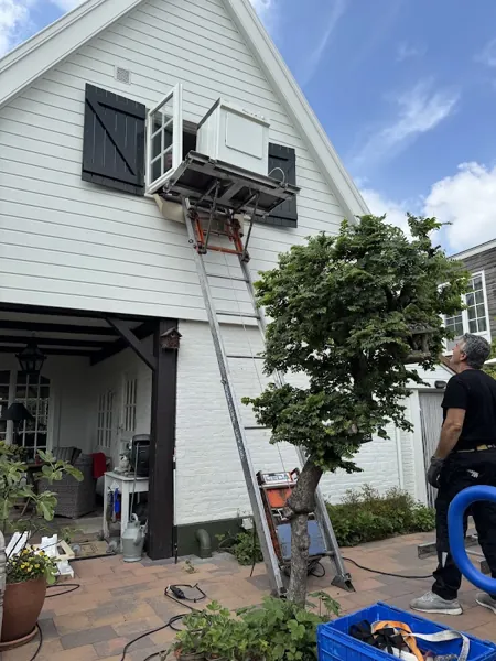 GEDA-lift tegen wit houten huis brengt witgoed naar een open raam met zwarte luiken. Bij Verhuislifthuren.nl huur je een verhuislift altijd met operator.