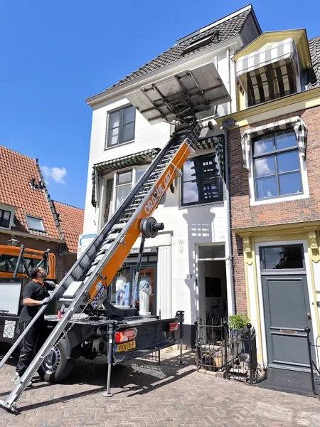 Vrachtwagen met ladderlift omhoog naar de zolder van een wit winkelpand met markiezen. Vandaag gebeld, morgen staat Verhuislifthuren.nl al voor je deur.